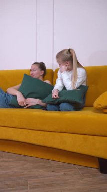 Anne ve kızı hardal sarısı bir kanepede oturup el ele tutuşarak endişeli ifadeler sergiliyorlar.