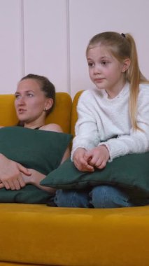 Anne ve kızı hardal sarısı bir kanepede oturup el ele tutuşarak endişeli ifadeler sergiliyorlar.