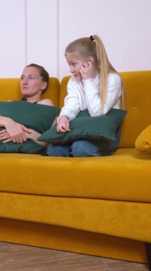 Anne ve kızı hardal sarısı bir kanepede oturup el ele tutuşarak endişeli ifadeler sergiliyorlar.