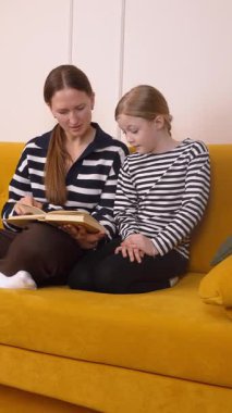 Genç okur yazarlığı beslemek, ilgili anne kızına rehberlik ederken yumuşak sarı koltukta birlikte okumak, rahat ev ortamında özel öğrenme deneyimini paylaşmak, dikey videolar.