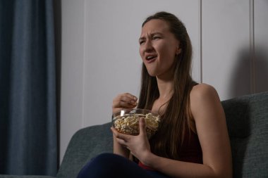 Gülümseyen genç kadın rahat bir koltukta oturup elinde cam kaseyle evde ilginç bir film izlerken patlamış mısır yemekten zevk alıyor.