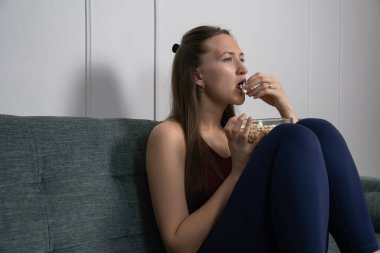 Kanepede rahat bir şekilde oturan genç bir kadın film izlerken cam kaseden patlamış mısırın tadını çıkarıyor. Evde huzurlu ve rahatlatıcı bir akşamın tadını çıkarıyor.