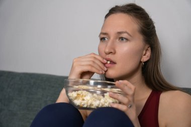 Kanepede rahat bir şekilde oturan genç bir kadın, film izlerken cam bir kaseden patlamış mısırın tadını çıkarıyor, evde boş zamanlarında tamamen dalmış ve anın tadını çıkarıyor.