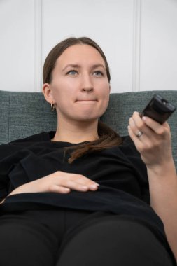 Genç bir kadın kanepeye uzanıp televizyon izliyor ve uzaktan kumandayla kanal değiştiriyor. Çeşitli programları ve programları izlerken sıkılmış bir yüz ifadesi sergiliyor. Dikey fotoğraf
