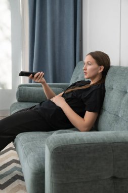 Comfortable young woman lounging on a sofa, watching television and holding a remote control, savoring a peaceful day at home filled with relaxation and leisure. Vertical photo