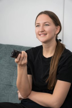 Young woman sitting comfortably on a sofa, watching television, using a remote control, changing channels and smiling, enjoying her leisure time at home. Vertical photo
