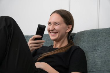 Young woman is lying on a comfortable sofa, holding a remote control and laughing while watching a funny tv show, enjoying her leisure time at home