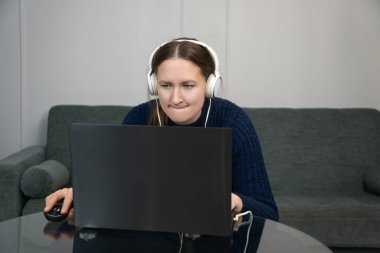 Kulaklık takan, dizüstü bilgisayarda çalışan ve ev ofisinin rahatlığıyla çalışırken ya da görevlerini tamamlarken odaklanmış bir ifadeyle ekrana bakan genç bir kadın.