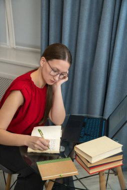 Kırmızı elbiseli genç bir kadın bir masada ders çalışıyor, dizüstü bilgisayar kullanıyor ve not defterine not alıyor. Kitaplar yakına istiflenmiş, verimli bir öğrenme ortamı yaratıyorlar. Dikey fotoğraf