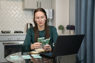 Genç bir kadın mutfak masasındaki avro banknotlarını titizlikle sayarken, online bankacılık ve bütçe işleri için dizüstü bilgisayar kullanıyor.