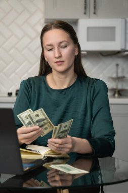 Genç iş kadını evinden işine odaklanıyor, mutfak masasında dizüstü bilgisayar ve defterle otururken internetten kazandığı paraları sayıyor, hesaplarını incelikle yönetiyor. Dikey fotoğraf
