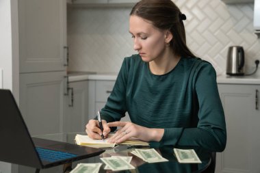 Not defterine notlar yazan, ev bütçesini yöneten, para hesaplayan ve mutfak masasında dizüstü bilgisayarla otururken finansal plan yapan bir kadın.