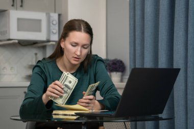 Ev bütçesini yöneten genç bir kadın dizüstü bilgisayar kullanırken dikkatle dolarları sayıyor. Muhtemelen online bankacılık ya da masraf takibi için.
