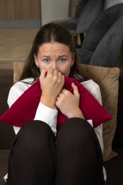 Stresli genç bir kadın kırmızı bir yastığı tutuyor ve gergin bir atmosferle çevrili parmaklarını endişeyle ısırıyor, iş yerinde endişe ve duygusal gerginliği somutlaştırıyor. Dikey fotoğraf