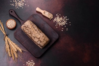 Tahta kesme tahtasının üzerinde tahıl taneleriyle birlikte koyu beton arka planda bir somun kahverengi ekmek.