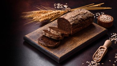 Tahta kesme tahtasının üzerinde tahıl taneleriyle birlikte koyu beton arka planda bir somun kahverengi ekmek.