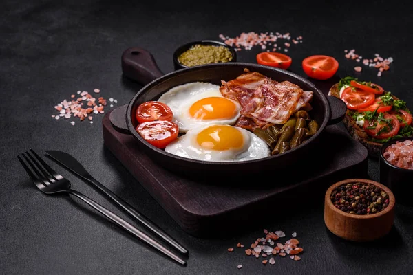 Stock image Tasty breakfast consists of eggs, bacon, beans, tomatoes, with spices and herbs on a dark concrete background