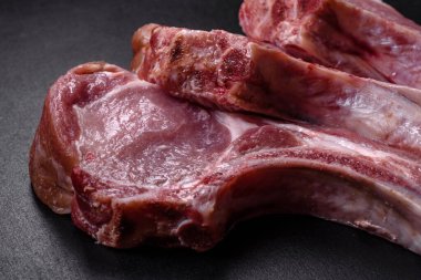 Kaburgalarda taze çiğ domuz eti, baharatlar ve ahşap kesme tahtasındaki otlar koyu beton arka planda.