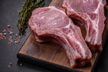 Kaburgalarda taze çiğ domuz eti, baharatlar ve ahşap kesme tahtasındaki otlar koyu beton arka planda.