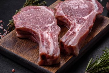 Kaburgalarda taze çiğ domuz eti, baharatlar ve ahşap kesme tahtasındaki otlar koyu beton arka planda.