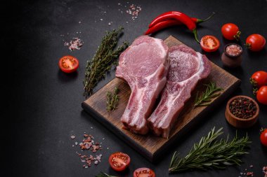 Kaburgalarda taze çiğ domuz eti, baharatlar ve ahşap kesme tahtasındaki otlar koyu beton arka planda.