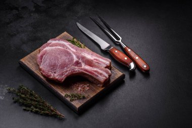 Kaburgalarda taze çiğ domuz eti, baharatlar ve ahşap kesme tahtasındaki otlar koyu beton arka planda.