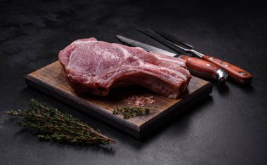 Kaburgalarda taze çiğ domuz eti, baharatlar ve ahşap kesme tahtasındaki otlar koyu beton arka planda.