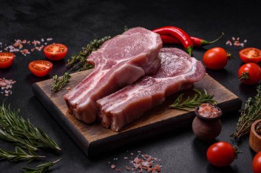 Kaburgalarda taze çiğ domuz eti, baharatlar ve ahşap kesme tahtasındaki otlar koyu beton arka planda.