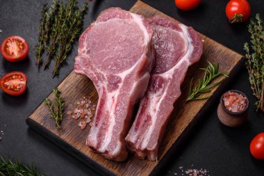 Kaburgalarda taze çiğ domuz eti, baharatlar ve ahşap kesme tahtasındaki otlar koyu beton arka planda.