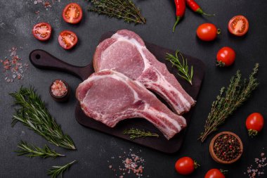 Kaburgalarda taze çiğ domuz eti, baharatlar ve ahşap kesme tahtasındaki otlar koyu beton arka planda.