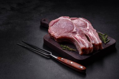 Kaburgalarda taze çiğ domuz eti, baharatlar ve ahşap kesme tahtasındaki otlar koyu beton arka planda.