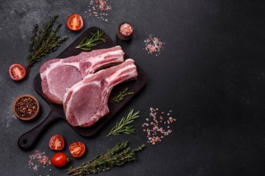 Kaburgalarda taze çiğ domuz eti, baharatlar ve ahşap kesme tahtasındaki otlar koyu beton arka planda.