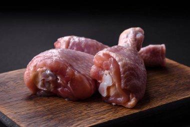 Three raw chicken legs with spices and herbs on a wooden cutting board against a dark concrete background