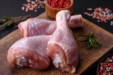 Three raw chicken legs with spices and herbs on a wooden cutting board against a dark concrete background