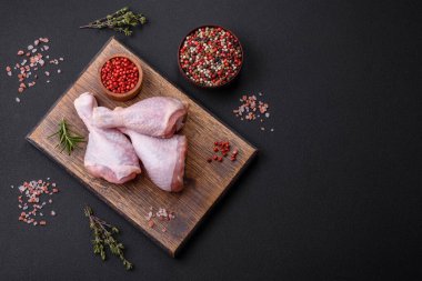 Three raw chicken legs with spices and herbs on a wooden cutting board against a dark concrete background