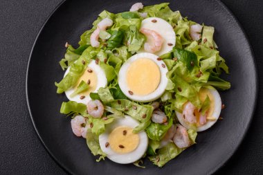 Delicious fresh healthy salad with shrimp, egg, lettuce and flax seeds on a black ceramic plate on a concrete background