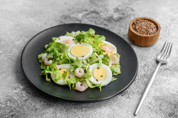Delicious fresh healthy salad with shrimp, egg, lettuce and flax seeds on a black ceramic plate on a concrete background
