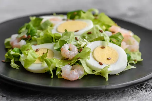Delicious fresh healthy salad with shrimp, egg, lettuce and flax seeds on a black ceramic plate on a concrete background