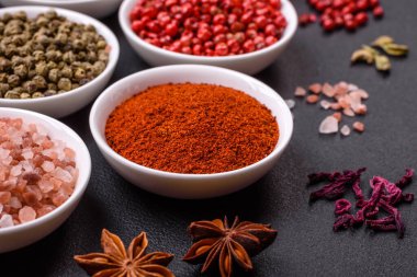 Composition consisting of variations of spices in white bowls and metal spoons on a dark concrete background