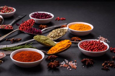 Composition consisting of variations of spices in white bowls and metal spoons on a dark concrete background