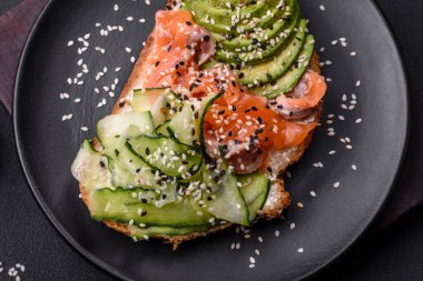 Fresh tasty sandwich with salmon, avocado and sesame and flax seeds on a dark concrete background