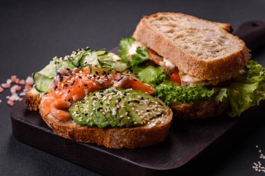 Fresh tasty sandwich with salmon, avocado and sesame and flax seeds on a dark concrete background