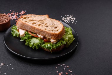 Fresh tasty sandwich with chicken, tomatoes and lettuce on a black plate on a dark concrete background