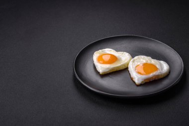 Two heart-shaped fried eggs on a black ceramic plate on a dark concrete background. Breakfast for valentine's day