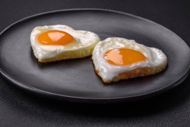 Two heart-shaped fried eggs on a black ceramic plate on a dark concrete background. Breakfast for valentine's day