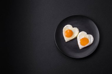 Two heart-shaped fried eggs on a black ceramic plate on a dark concrete background. Breakfast for valentine's day
