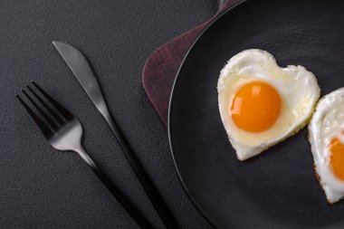Two heart-shaped fried eggs on a black ceramic plate on a dark concrete background. Breakfast for valentine's day