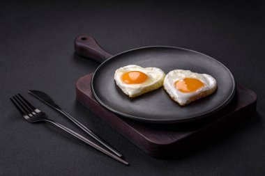 Two heart-shaped fried eggs on a black ceramic plate on a dark concrete background. Breakfast for valentine's day