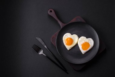 Two heart-shaped fried eggs on a black ceramic plate on a dark concrete background. Breakfast for valentine's day