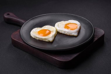 Two heart-shaped fried eggs on a black ceramic plate on a dark concrete background. Breakfast for valentine's day